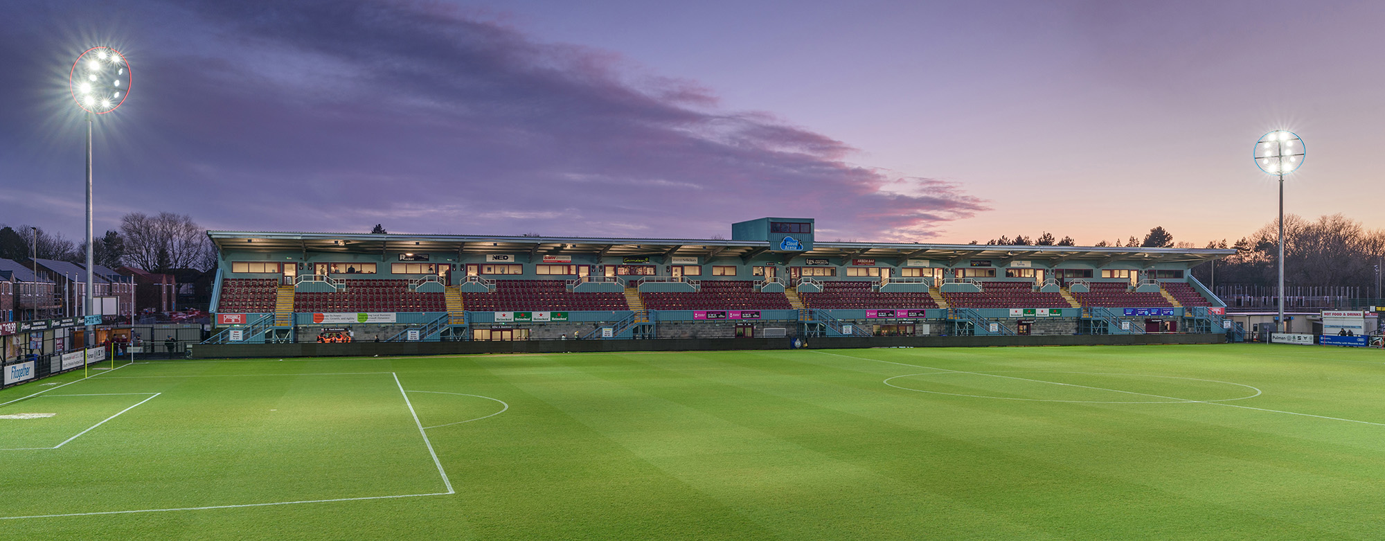 Wide exterior view of a football pitch with main stand at dusk with floodlights and dramatic sky
