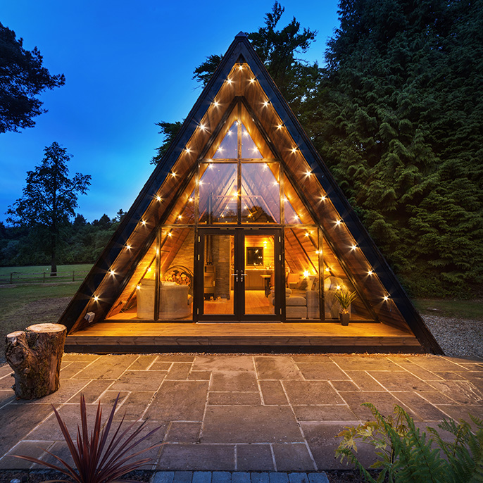 An A-frame garden building illuminated at dusk with large trees in the landscape behind and in the distance