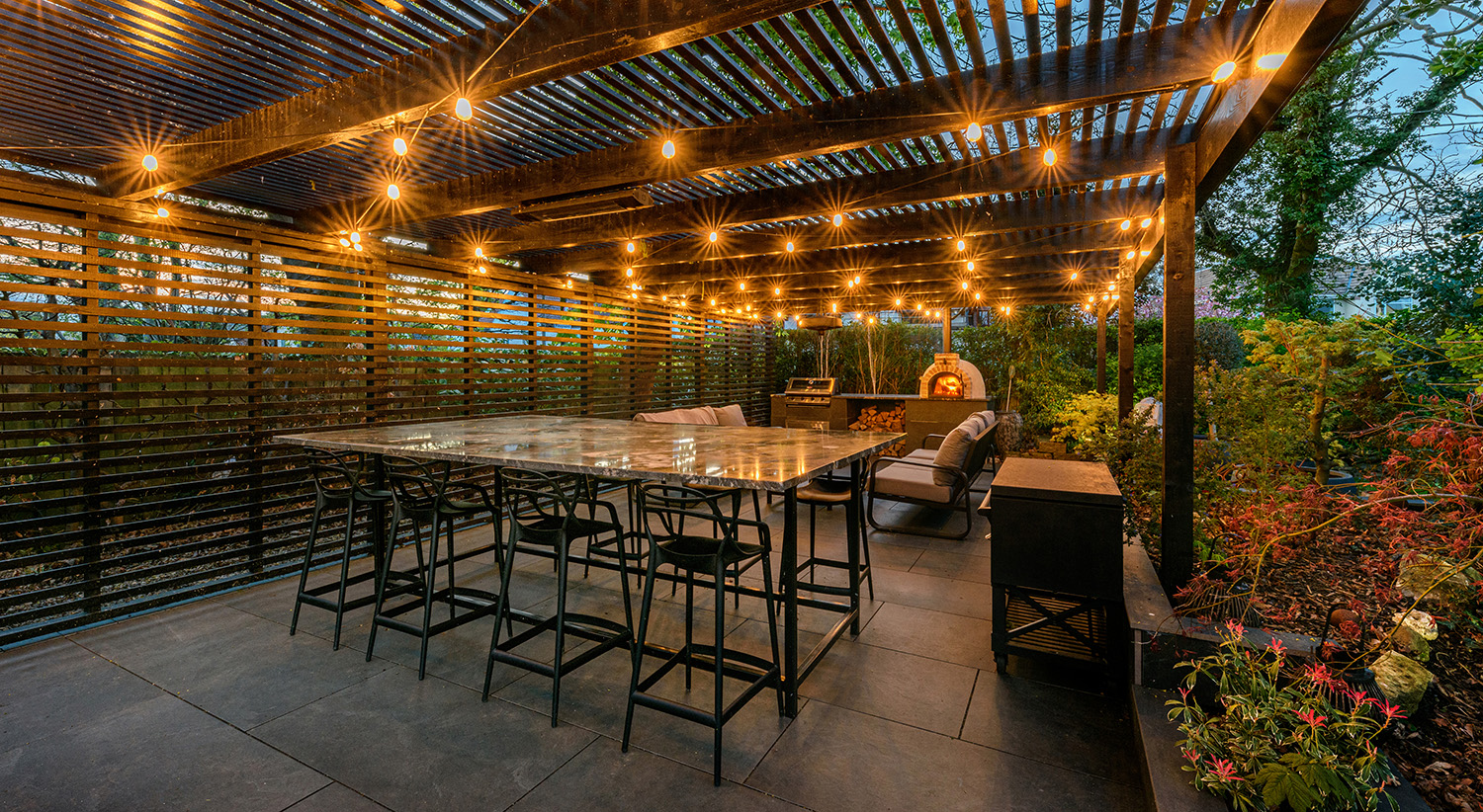 Garden dining area at dusk with lighting and pizza oven
