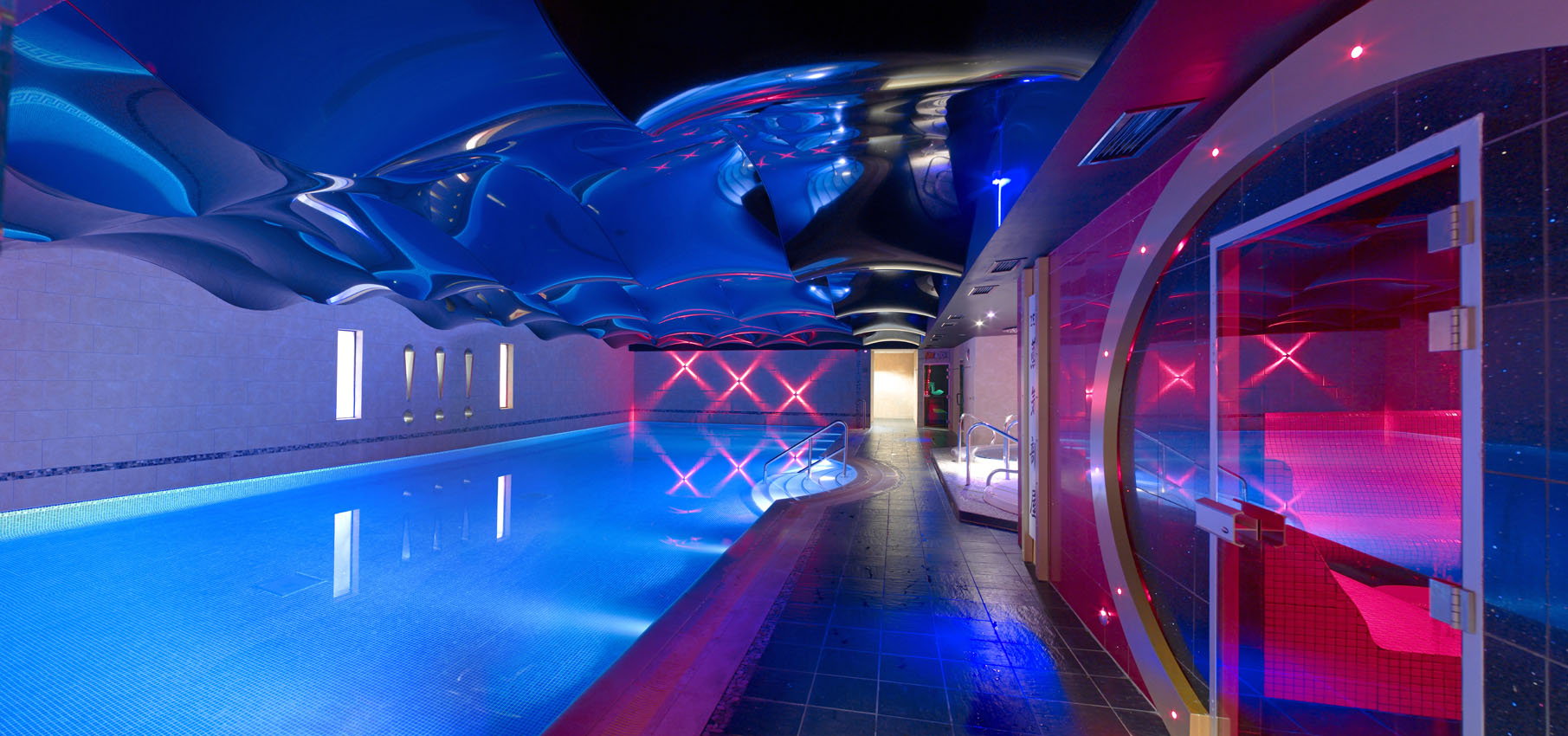 Illuminated modern indoor swimming pool in with mirrored ceiling and blue and pink feature lighting